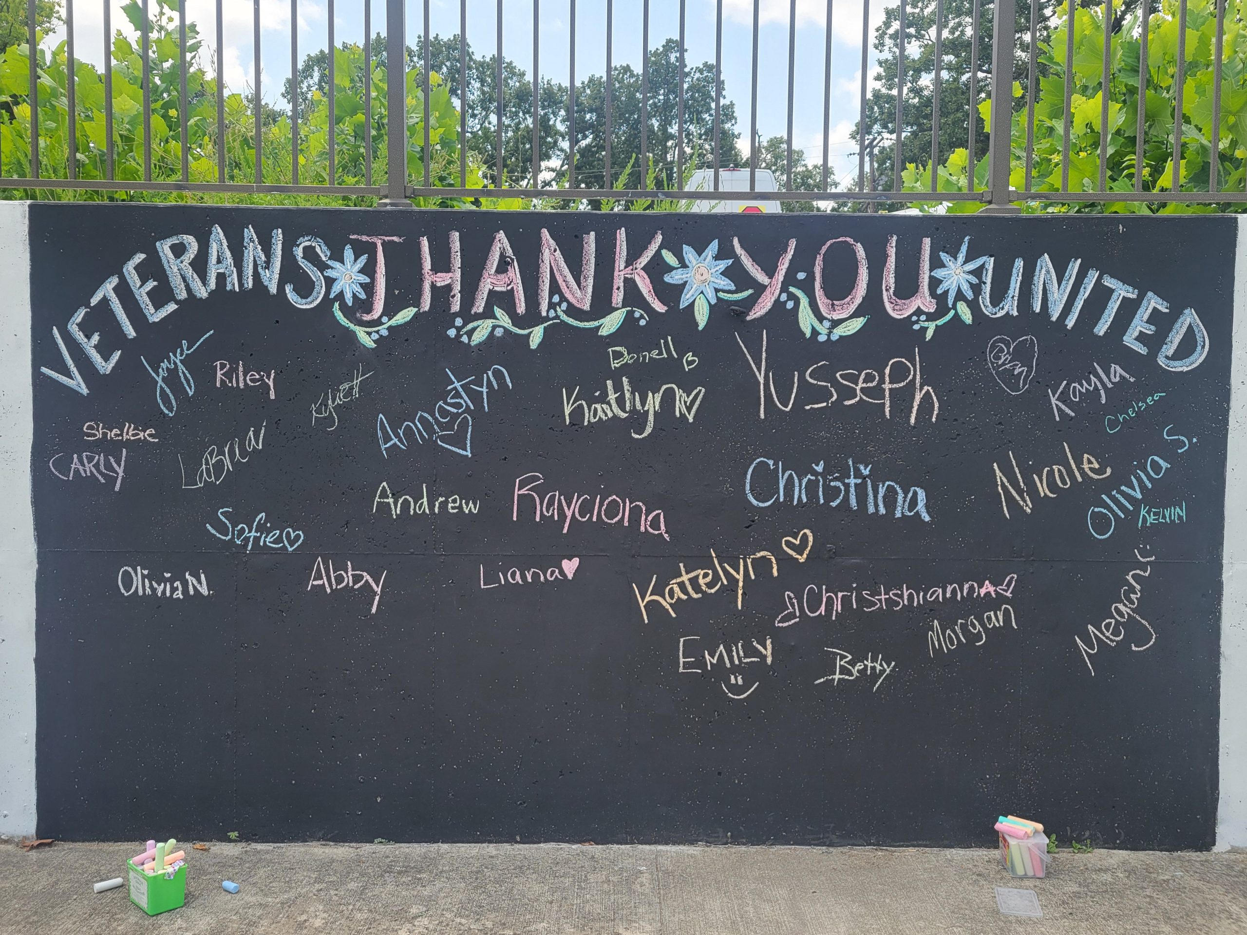 A dark chalkboard banner displayed on a computer screen featuring the handwritten message "VETERANS THANK YOU UNITED" in large, stylized white capital letters. The banner is filled with numerous individual names and signatures handwritten in white chalk, some accompanied by small drawings like hearts.